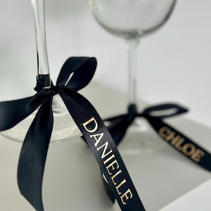 May include: Two wine glasses with black ribbon bows and gold lettering. The ribbons are tied around the stems of the glasses and have the names "Danielle" and "Chloe" written on them.