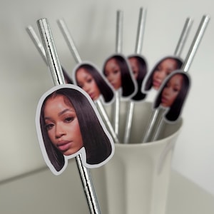 May include: A collection of silver paper straws, each adorned with a sticker featuring a woman's face. The straws are arranged in a white ceramic cup, creating a decorative and personalized touch. The stickers show a woman with long, dark hair.