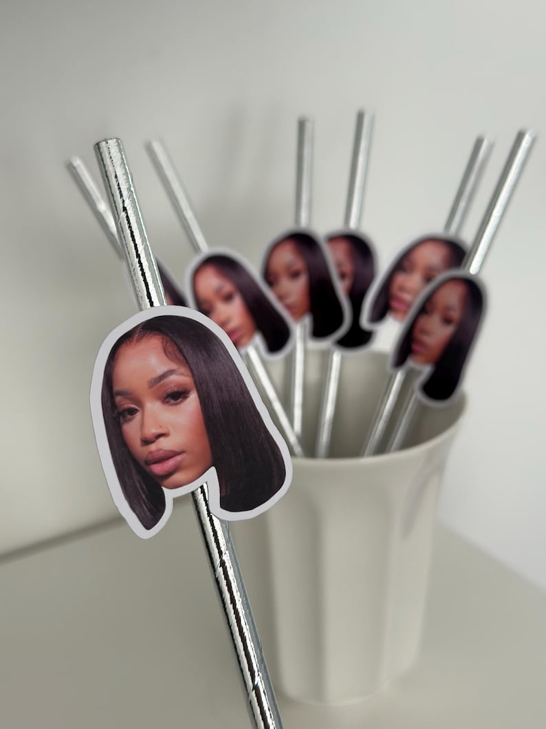 May include: A collection of silver paper straws, each adorned with a sticker featuring a woman's face. The straws are arranged in a white ceramic cup, creating a decorative and personalized touch. The stickers show a woman with long, dark hair.