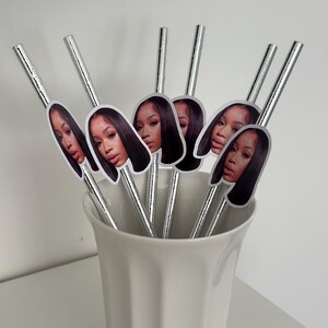 May include: A collection of silver-colored drinking straws, each adorned with a sticker featuring a woman's face. The straws are arranged in a white ceramic cup, creating a decorative and stylish presentation. The image is well-lit, highlighting the details of the straws and stickers.
