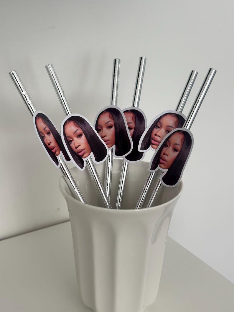 May include: A collection of silver-colored drinking straws, each adorned with a sticker featuring a woman's face. The straws are arranged in a white ceramic cup, creating a decorative and stylish presentation. The image is well-lit, highlighting the details of the straws and stickers.