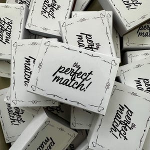 Może przedstawiać: Stos małych, białych pudełek z napisem "the perfect match!" czarnym skryptem. Każde pudełko ozdobione jest delikatną czarną obwódką z serc i zawijasów. Pudełka są ułożone w stos.