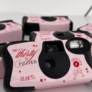 May include: Pink disposable cameras with black accents. The cameras have text that reads "Chanel at 30", "thirty", "Viva Mexico", and "30.08.95". The cameras also have red star and wine glass graphics.