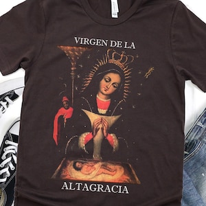 Peut inclure: Un t-shirt marron avec une image vintage de la Vierge Marie et de l'enfant Jésus. L'image est entourée d'un cadre doré et du texte "VIRGEN DE LA ALTAGRACIA".