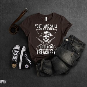 May include: Brown t-shirt with a white skull and crossbones graphic. The text reads "Youth and skill are no match for old age and treachery".