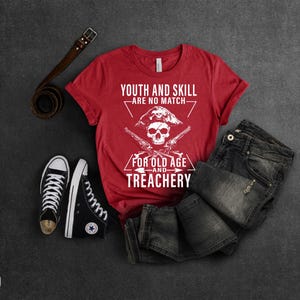 May include: Red t-shirt with a white skull and crossbones graphic. The text on the shirt reads "Youth and skill are no match for old age and treachery".