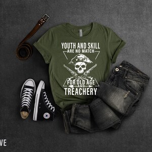 May include: Olive green t-shirt with a white skull and crossbones graphic. The graphic says "Youth and Skill Are No Match For Old Age and Treachery".