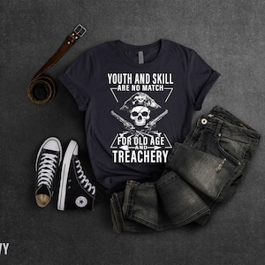 May include: A navy blue t-shirt with a white skull and crossbones graphic. The text on the shirt reads "Youth and Skill - Are No Match - For Old Age and Treachery".
