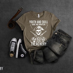 May include: A brown t-shirt with a white graphic design featuring a skull with crossed pistols and the text "Youth and Skill Are No Match For Old Age and Treachery".