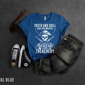 May include: Royal blue t-shirt with a white skull and crossbones graphic. The text on the shirt reads "Youth and Skill Are No Match For Old Age and Treachery".