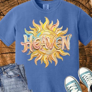 May include: A blue t-shirt with a textured sun design and the word "HEAVEN". The sun is in yellow, orange, and pink. The shirt is paired with blue jeans and teal trainers.
