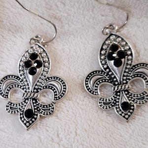 May include: A pair of silver-toned fleur-de-lis earrings. Each earring features a detailed design with black gemstone accents. The earrings have a hook closure and are displayed on a textured, off-white surface.