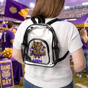 May include: A clear plastic backpack with black trim and black mesh side pockets. The front pocket features a graphic of a tiger face above the word Tigers in purple and gold lettering, with a purple bow and the name ANN printed in purple below.