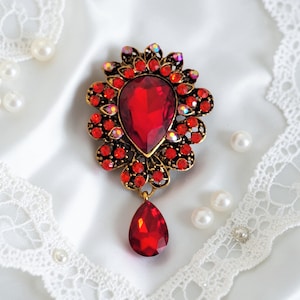 May include: A vintage-style brooch with a large, teardrop-shaped red gemstone pendant. The brooch features a gold-toned metal setting with smaller red and iridescent crystals. The design is floral, with a central red stone and a dangling teardrop.