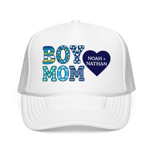 May include: A white trucker hat with a mesh back and a front graphic featuring the text BOY MOM in blue and teal patterned letters, accompanied by a dark blue heart shape containing the white text NOAH + NATHAN printed on the front panel.