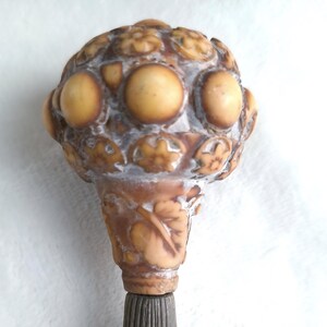 May include: A detailed close-up of a decorative cane handle. The handle features a rounded top with a pattern of raised circles and floral designs in shades of tan and brown. The lower portion has a leaf design, and the base is a dark, textured material.