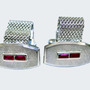 May include: A pair of silver cufflinks with a textured surface and two rectangular red gemstones. The cufflinks have a mesh chain design for the closure, and are set against a light background.