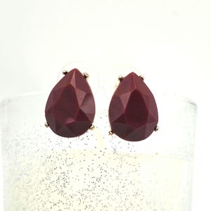 May include: A pair of teardrop-shaped stud earrings. The earrings feature a faceted, burgundy-colored stone set in a gold-tone metal. The earrings are displayed against a white background.