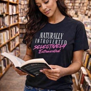 May include: Black t-shirt with the text "NATURALLY INTROVERTED SELECTIVELY Extroverted" in white and pink. The person is in a library, holding a book and wearing blue jeans.
