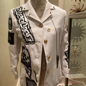 Puede incluir: Una chaqueta blanca con diseños gráficos en blanco y negro, incluyendo la palabra "DISCLOSE" y una calavera. La chaqueta tiene botones dorados y un detalle de cadena.