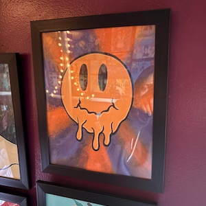 Op de afbeelding: Ingelijst kunstwerk met een levendig oranje smiley-gezicht met een smeltend ontwerp. Het kunstwerk is geplaatst tegen een achtergrond van oranje en paarse tinten, omlijst in een zwart frame. Het kunstwerk wordt tentoongesteld op een bordeauxrode muur.