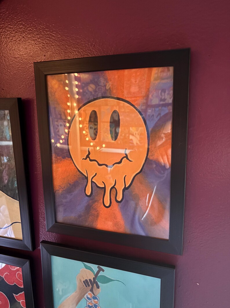 Op de afbeelding: Ingelijst kunstwerk met een levendig oranje smiley-gezicht met een smeltend ontwerp. Het kunstwerk is geplaatst tegen een achtergrond van oranje en paarse tinten, omlijst in een zwart frame. Het kunstwerk wordt tentoongesteld op een bordeauxrode muur.