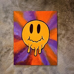 Op de afbeelding: Een canvas schilderij met een gele, smeltende smiley tegen een levendige achtergrond. De achtergrond heeft een radiaal ontwerp met oranje en paarse tinten. De smiley heeft zwarte ovale ogen en een druipende mond.