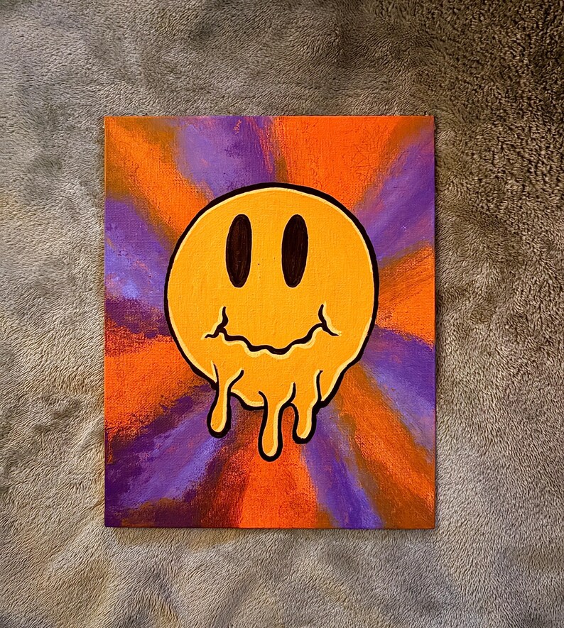 Op de afbeelding: Een canvas schilderij met een gele, smeltende smiley tegen een levendige achtergrond. De achtergrond heeft een radiaal ontwerp met oranje en paarse tinten. De smiley heeft zwarte ovale ogen en een druipende mond.