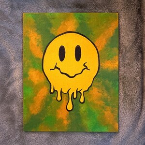 Op de afbeelding: Een canvas schilderij met een gele, smeltende smiley met zwarte contouren tegen een groene en oranje achtergrond. Het kunstwerk heeft een psychedelische, retro vibe.