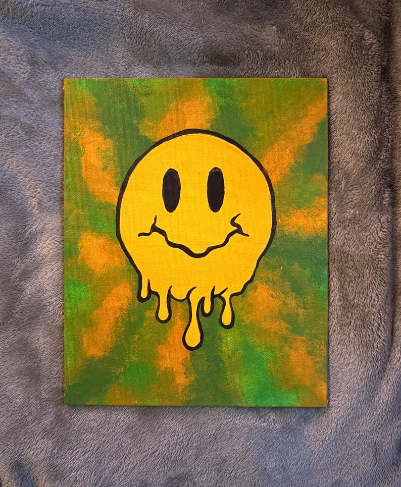 Op de afbeelding: Een canvas schilderij met een gele, smeltende smiley met zwarte contouren tegen een groene en oranje achtergrond. Het kunstwerk heeft een psychedelische, retro vibe.