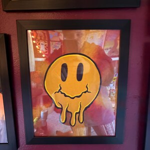 Op de afbeelding: Een ingelijst kunstwerk met een gele, smeltende smiley met zwarte ogen en mond. Het kunstwerk zit in een zwarte lijst en hangt aan een bordeauxrode muur. Andere ingelijste kunstwerken zijn zichtbaar.
