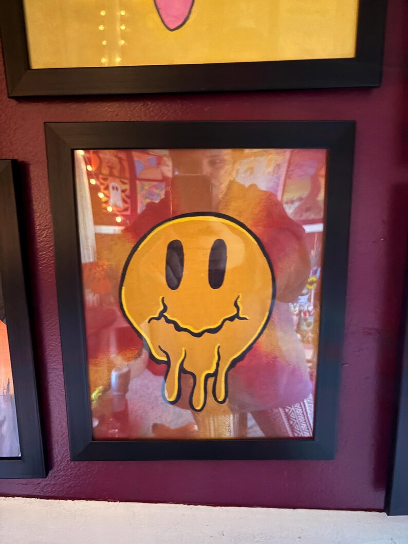 Op de afbeelding: Een ingelijst kunstwerk met een gele, smeltende smiley met zwarte ogen en mond. Het kunstwerk zit in een zwarte lijst en hangt aan een bordeauxrode muur. Andere ingelijste kunstwerken zijn zichtbaar.