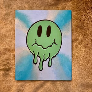 Op de afbeelding: Een canvas schilderij met een groene, smeltende smiley met zwarte ogen en een zwarte omtrek. De achtergrond heeft een blauw-wit tie-dye effect. Het kunstwerk wordt getoond op een bruin oppervlak.