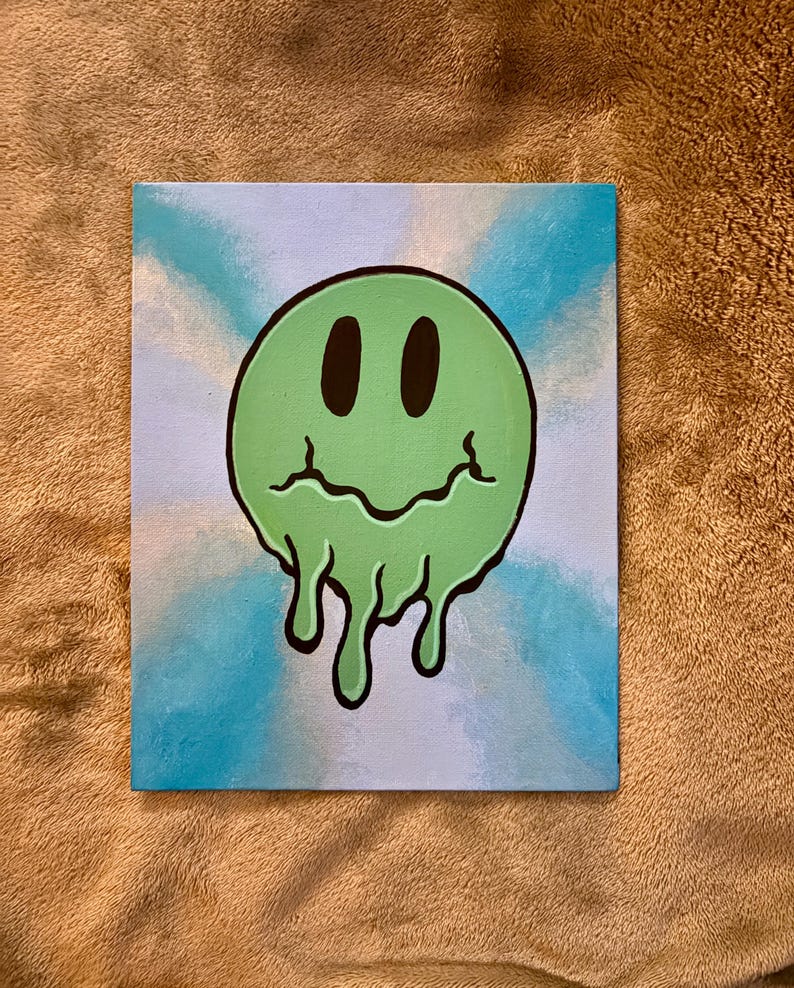 Op de afbeelding: Een canvas schilderij met een groene, smeltende smiley met zwarte ogen en een zwarte omtrek. De achtergrond heeft een blauw-wit tie-dye effect. Het kunstwerk wordt getoond op een bruin oppervlak.