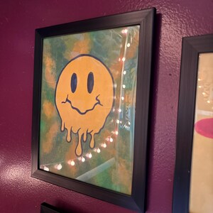 Op de afbeelding: Ingelijst kunstwerk met een gele smiley met een smeltend ontwerp. Het kunstwerk heeft zwarte ogen en een gebogen mond. De achtergrond is een mix van groen en geel. De lijst is zwart.