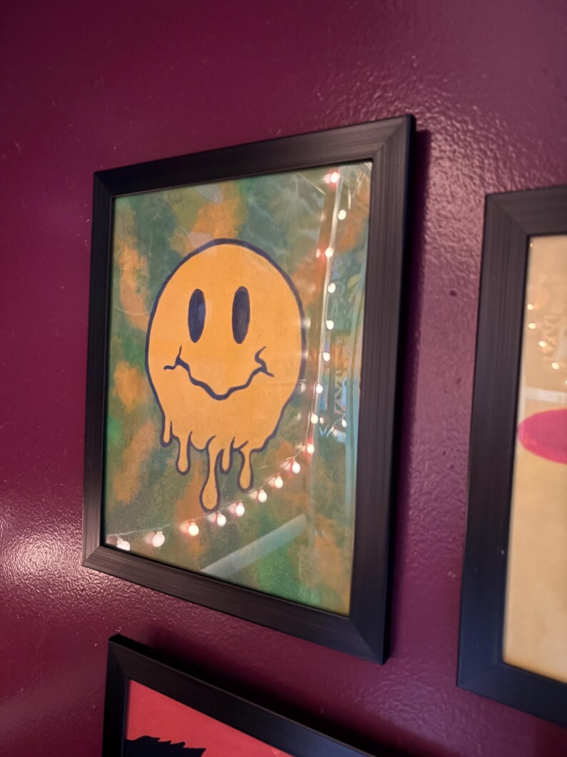 Op de afbeelding: Ingelijst kunstwerk met een gele smiley met een smeltend ontwerp. Het kunstwerk heeft zwarte ogen en een gebogen mond. De achtergrond is een mix van groen en geel. De lijst is zwart.