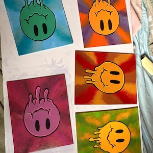 Op de afbeelding: Vijf vierkante canvas schilderijen met een gestileerd smiley gezicht. Elk gezicht heeft een andere kleur, waaronder groen, oranje, roze, geel en oranje, met zwarte ovale ogen. De achtergronden zijn een mix van kleuren, waaronder blauw, paars, rood en groen.