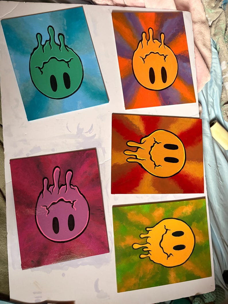 Op de afbeelding: Vijf vierkante canvas schilderijen met een gestileerd smiley gezicht. Elk gezicht heeft een andere kleur, waaronder groen, oranje, roze, geel en oranje, met zwarte ovale ogen. De achtergronden zijn een mix van kleuren, waaronder blauw, paars, rood en groen.