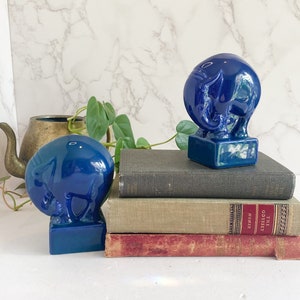 May include: Two cobalt blue ceramic elephant figurines, each mounted on a rectangular base. The elephants are placed on a stack of vintage books with a brass teapot and greenery in the background. The figurines are glossy and reflect light.
