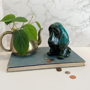 May include: A vintage brass teapot holds a green leafy plant. A ceramic, dark teal-colored basset hound figurine sits on a blue book. Several coins are scattered near the book, creating a still-life composition.