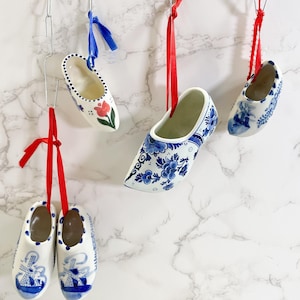May include: Several white ceramic clogs with blue floral and windmill designs are suspended by red and blue ribbons. One clog features a red tulip. The clogs are hanging against a white marble background.