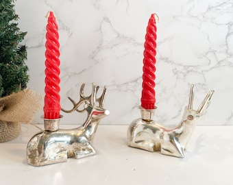 May include: Two silver-colored reindeer candle holders with red spiral candles. A small green Christmas tree is in the background. The candle holders are on a white surface with a marble-patterned background.