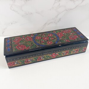 May include: A rectangular wooden box with a hinged lid. The box is painted black with floral designs in red, green, and blue. The lid features a central floral pattern within a blue and gold border. The sides of the box are decorated with a repeating floral motif.
