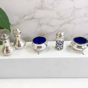 May include: A collection of antique silver salt and pepper shakers and salt cellars. The shakers have a classic pear shape with perforated tops. The salt cellars have a silver base with a deep blue glass interior. The salt cellars are supported by three small feet.