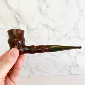 May include: A brown wooden pipe with intricate carvings and a black mouthpiece. The pipe is held in a hand.