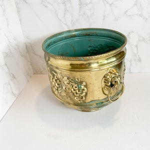May include: A gold planter with a green interior and grapevine design. The planter has two lion head handles with rings.