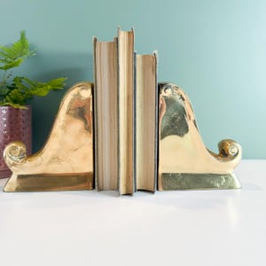 May include: A pair of brass bookends with a curved design, holding several books with aged pages. The bookends have a polished, reflective surface. A potted plant is visible in the background.
