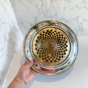May include: Silver and gold metal ashtray with a decorative, geometric design. The ashtray has a round shape and a raised rim.