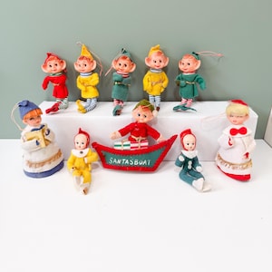 May include: A collection of ten vintage-style felt elf ornaments with painted faces. The figures include elves in red, yellow, green, and white outfits, some holding books or gifts. One elf sits in a red felt boat labeled SANTASBOAT in white text.