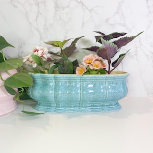 May include: A light blue ceramic planter filled with pink and yellow flowers and green and purple foliage. The planter has a textured surface and a scalloped design. The planter is on a white surface.